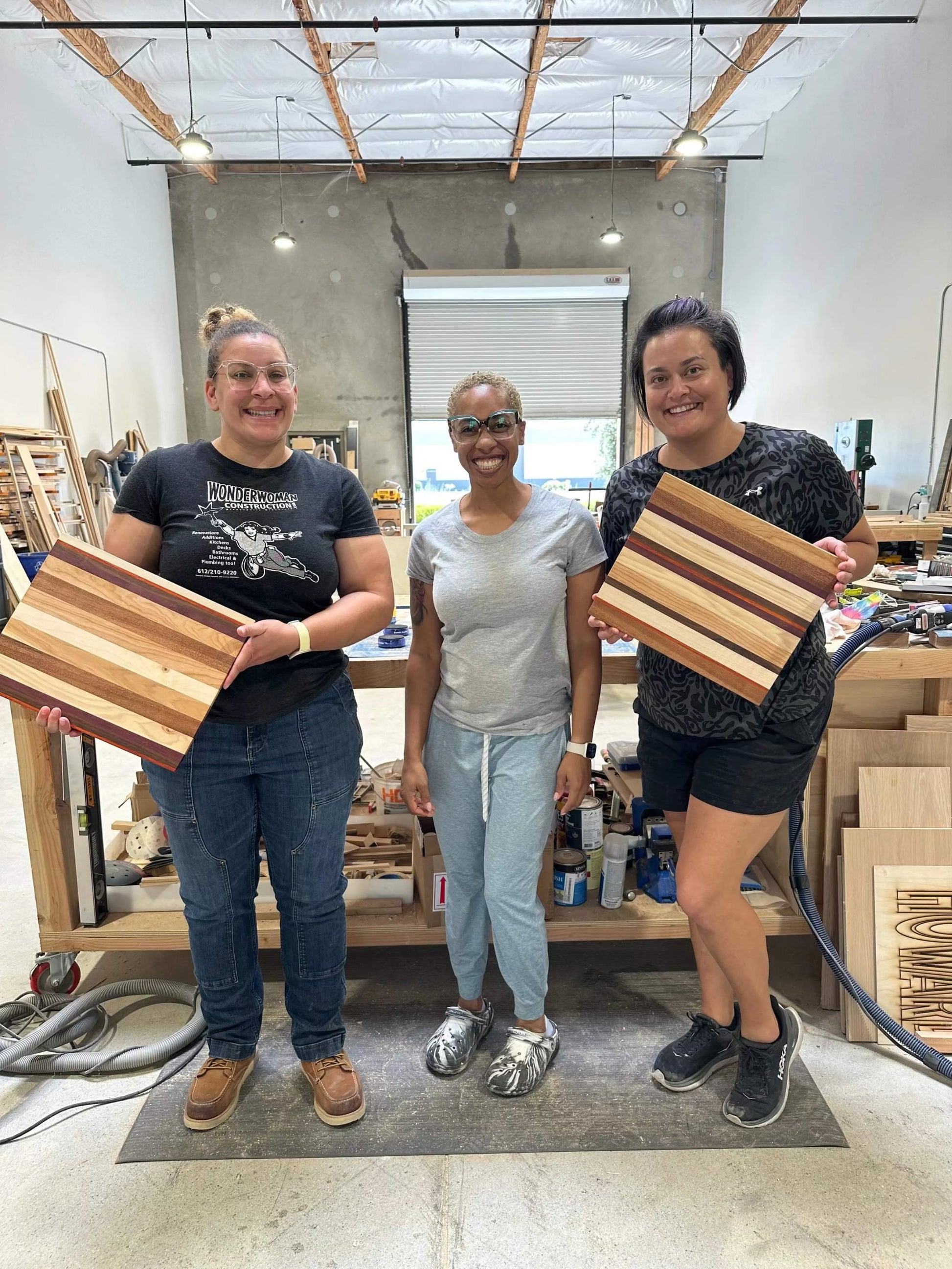 Three women holding wooden cutting boards in a workshop setting.