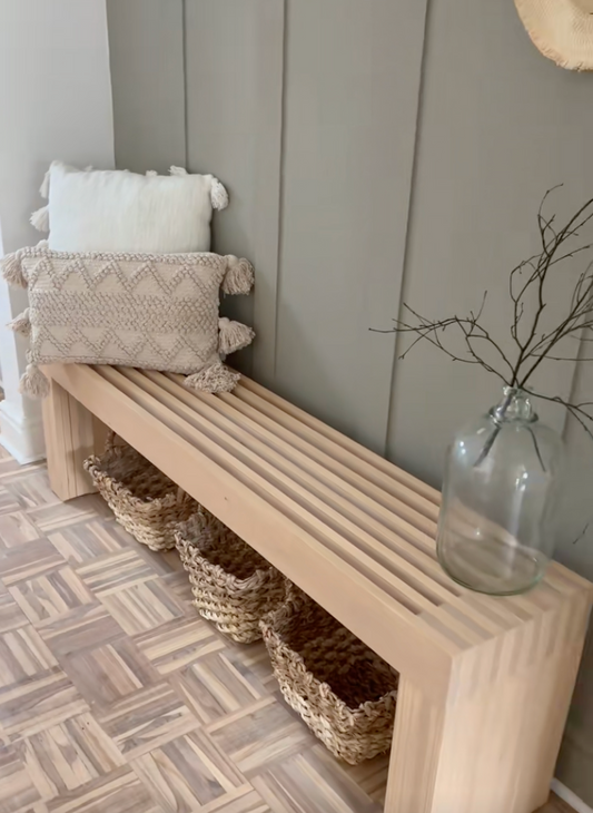 Modern wooden slatted bench styled in an entryway with neutral throw pillows, woven baskets underneath for storage, and a glass vase with decorative branches. Light natural wood bench placed against a vertical paneled wall with warm, minimalist home decor.