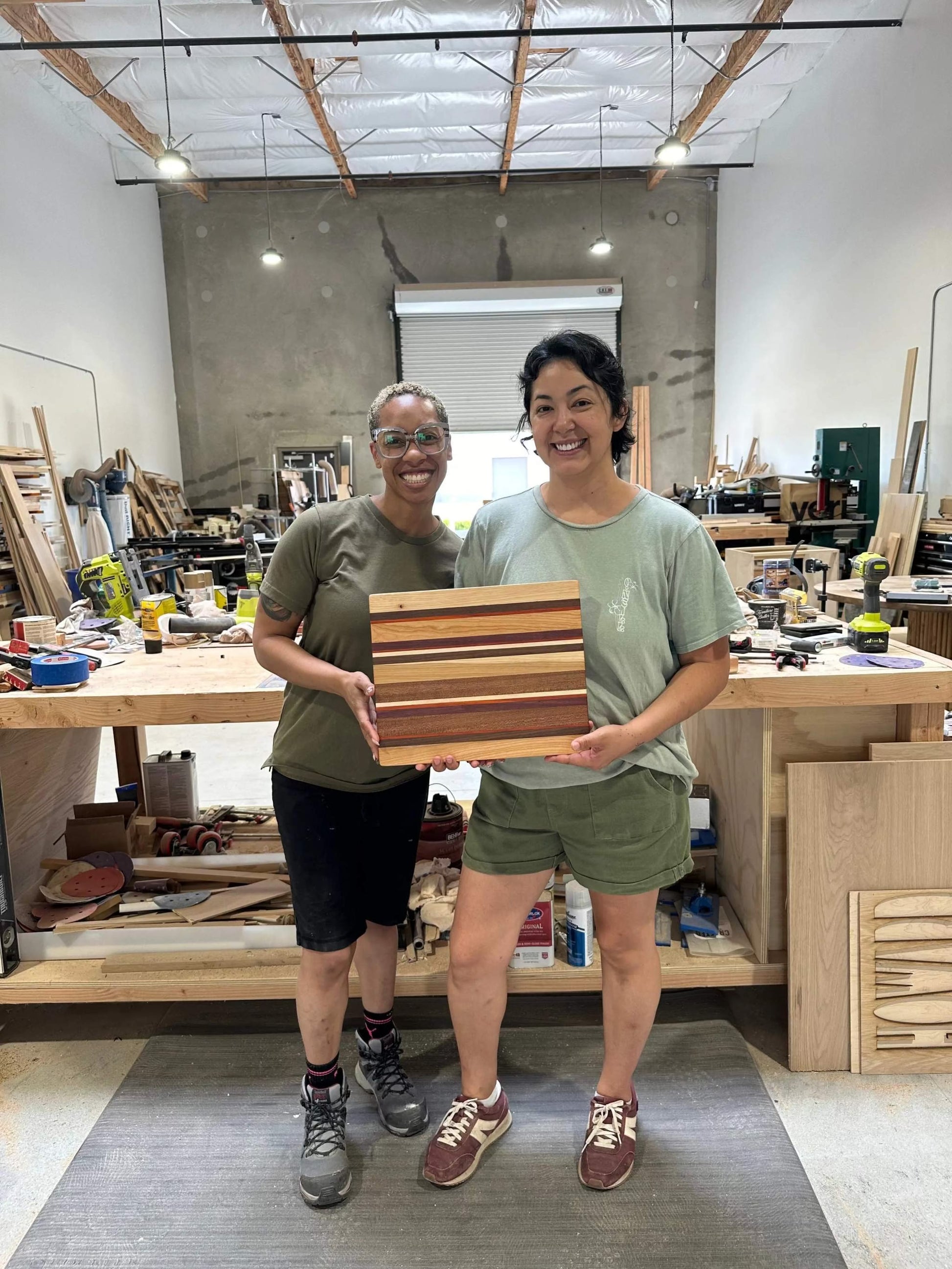 Two people holding a wooden cutting board in a workshop setting.