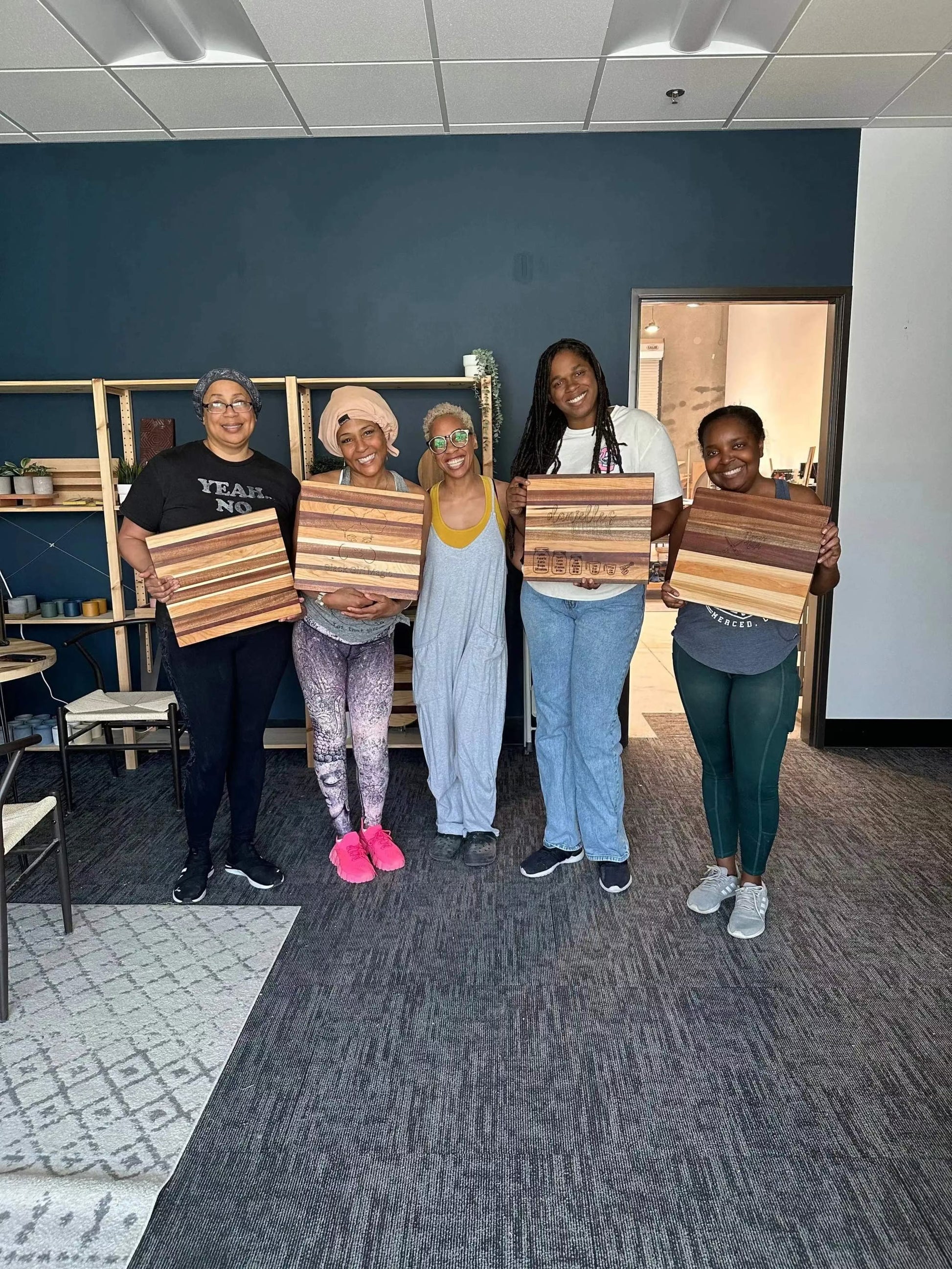 Five smiling students holding handmade striped wooden cutting boards during a woodworking class inside a workshop. Participants proudly display their finished boards made from mixed hardwoods, showcasing custom craftsmanship and beginner-friendly woodworking experience.