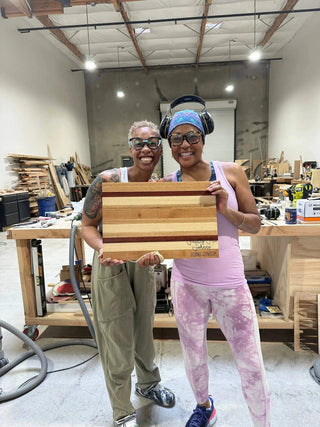 Two people holding a wooden cutting board in a workshop setting.